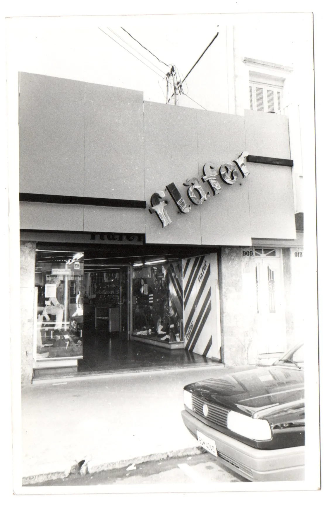 Fotografia em preto e branco da fachada de uma das primeiras lojas Flafer, com o nome em letras aplicadas sobre a parede, vitrine iluminada e um carro estacionado em frente